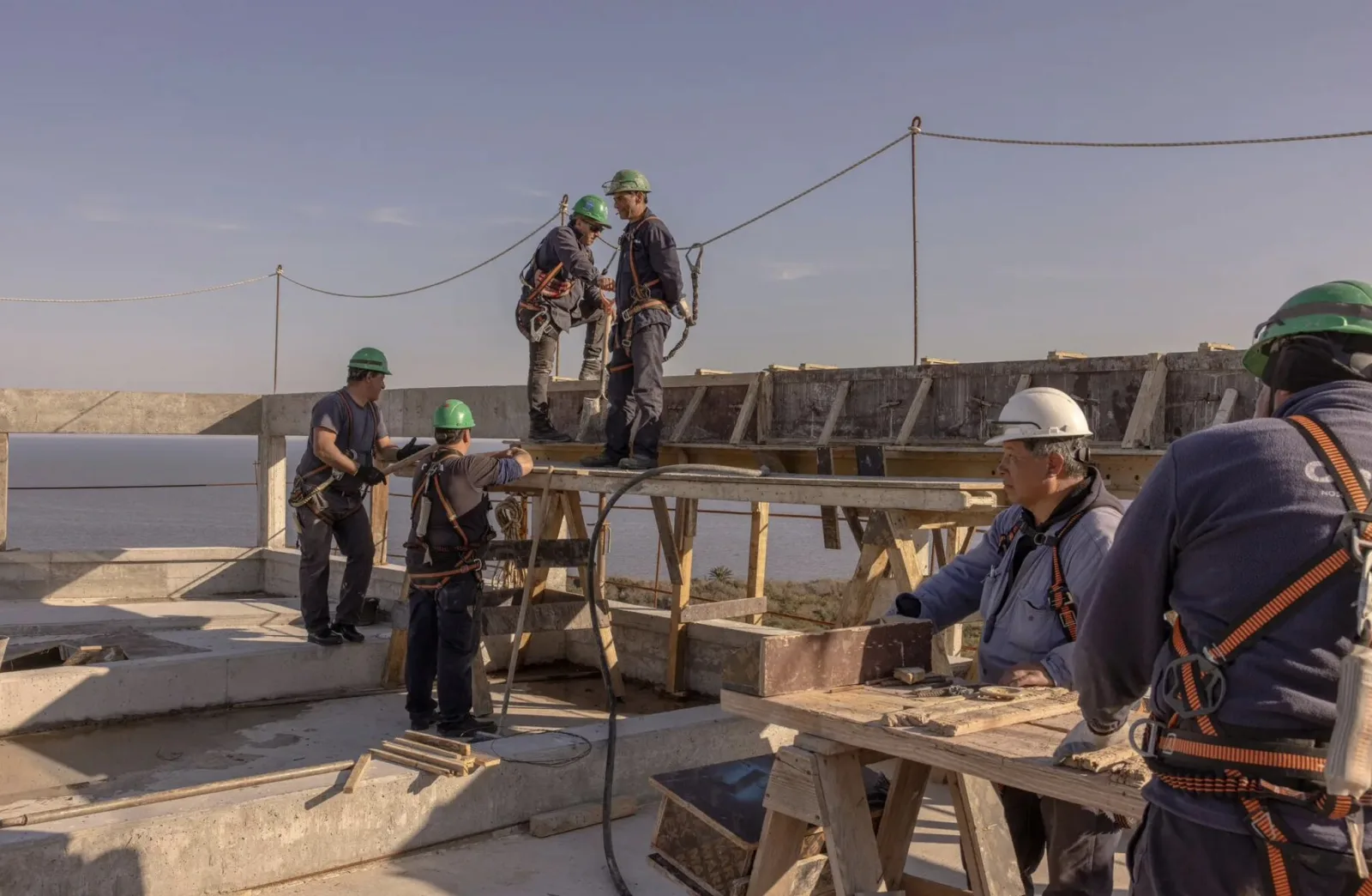 Construction crew in harnesses and hard hats setting formwork on a rooftop above the Río de la Plata.